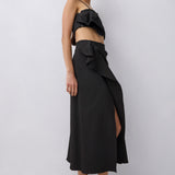 A model wearing a black ruffled two-piece set consisting of a strapless crop top and a high-waisted wrap maxi skirt with a side slit, paired with black strappy mule sandals, against a light gray studio background.