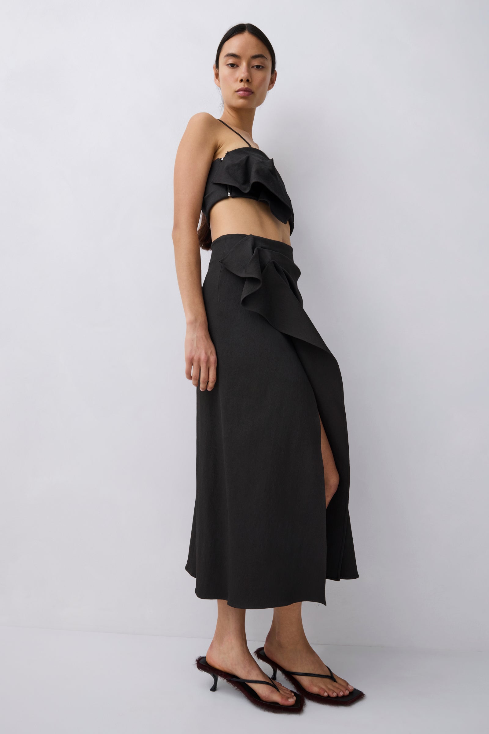 A model wearing a black ruffled two-piece set consisting of a strapless crop top and a high-waisted wrap maxi skirt with a side slit, paired with black strappy mule sandals, against a light gray studio background.