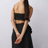 A woman shown from behind wearing a black two-piece outfit consisting of a strapless wrap-style crop top and a long skirt, against a plain light gray studio background.