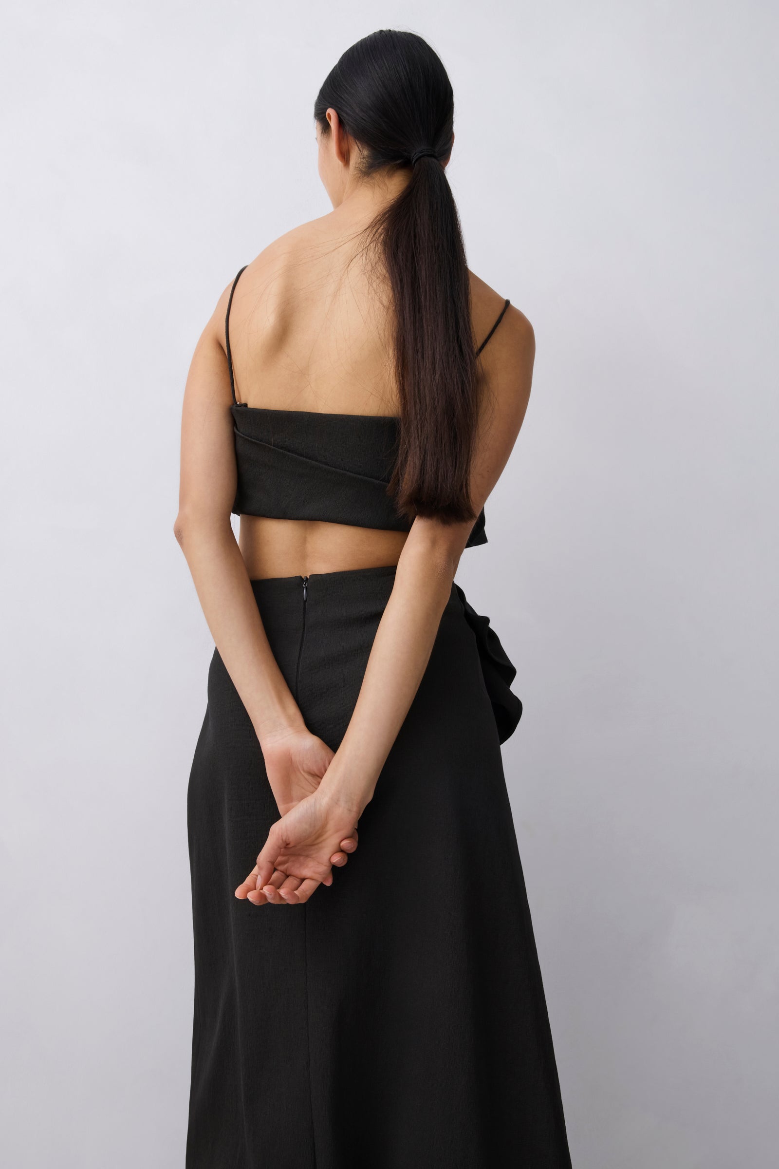 A woman shown from behind wearing a black two-piece outfit consisting of a strapless wrap-style crop top and a long skirt, against a plain light gray studio background.