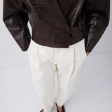 A cropped studio shot of a model wearing a dark brown crocodile-embossed wrap jacket, white pleated trousers, and dark brown pointed-toe flats against a light gray background.