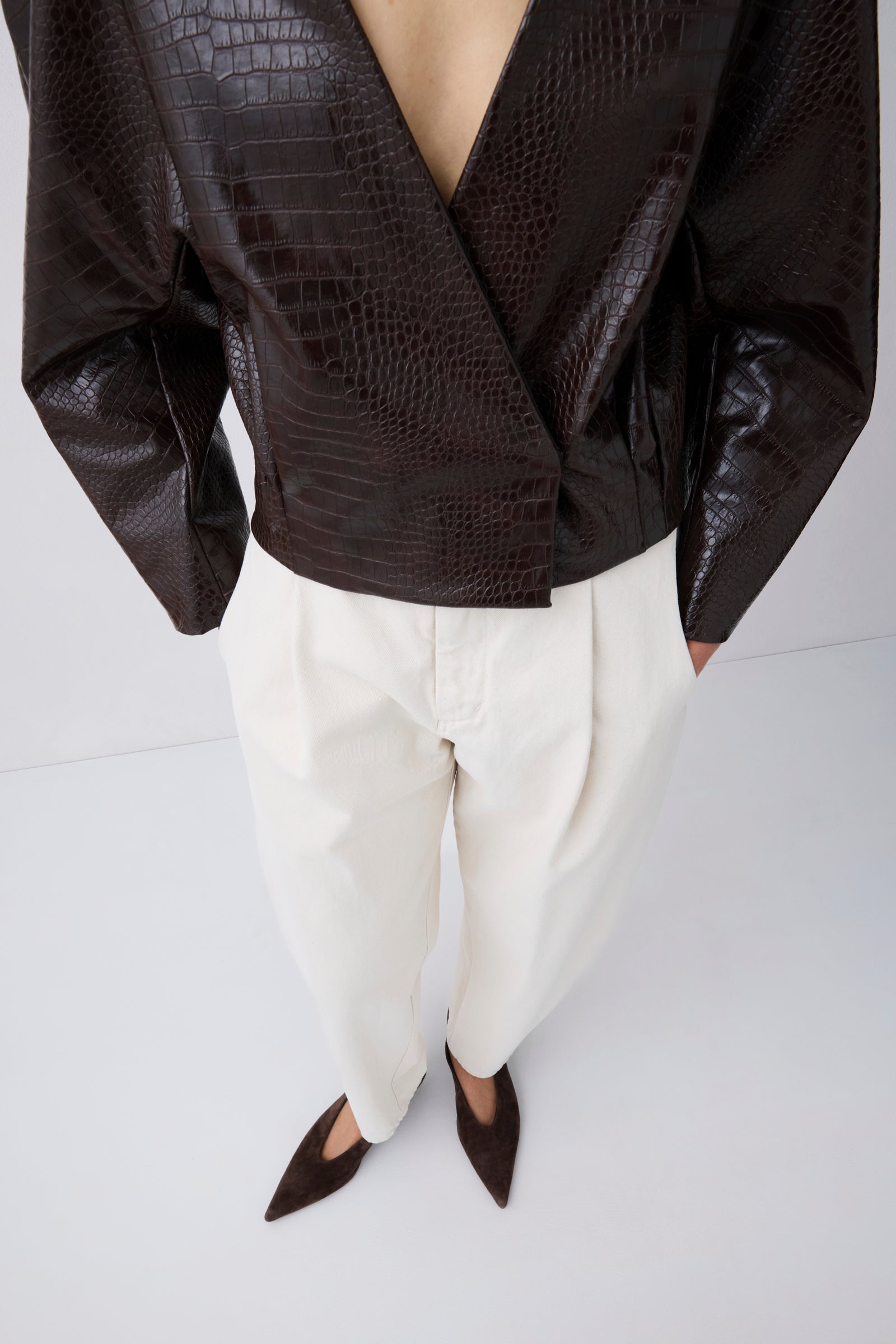 A cropped studio shot of a model wearing a dark brown crocodile-embossed wrap jacket, white pleated trousers, and dark brown pointed-toe flats against a light gray background.