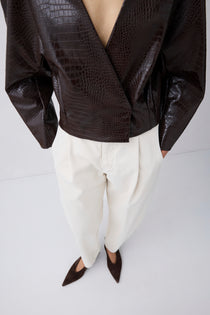 A cropped studio shot of a model wearing a dark brown crocodile-embossed wrap jacket, white pleated trousers, and dark brown pointed-toe flats against a light gray background.
