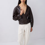 A model wearing a dark brown textured faux-leather wrap top with long sleeves, paired with off-white wide-leg trousers and dark brown pointed flats, standing against a light gray studio background.