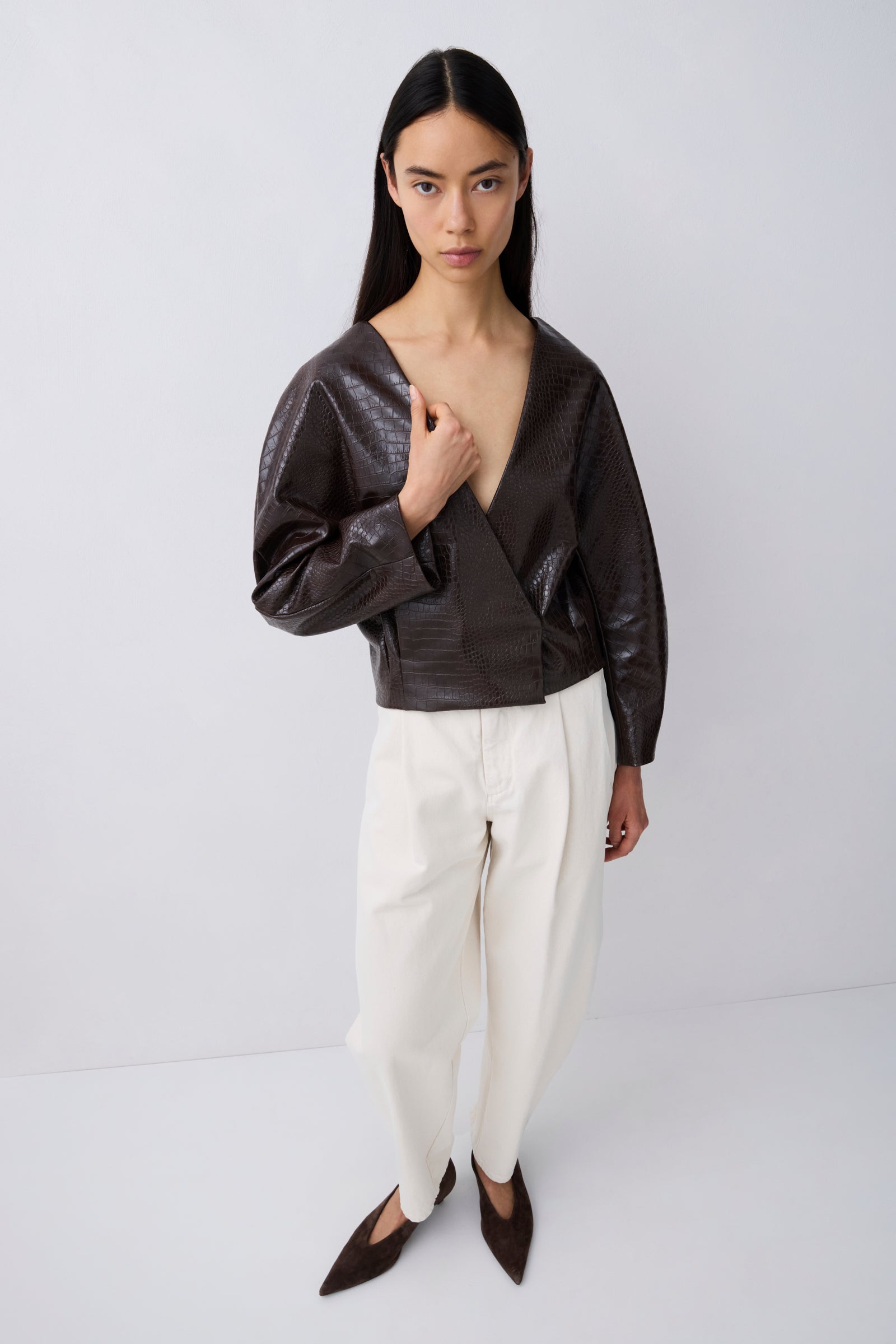 A model wearing a dark brown textured faux-leather wrap top with long sleeves, paired with off-white wide-leg trousers and dark brown pointed flats, standing against a light gray studio background.