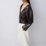 A model wearing a dark brown textured faux-leather wrap jacket and cream wide-leg trousers, standing against a light gray studio background.