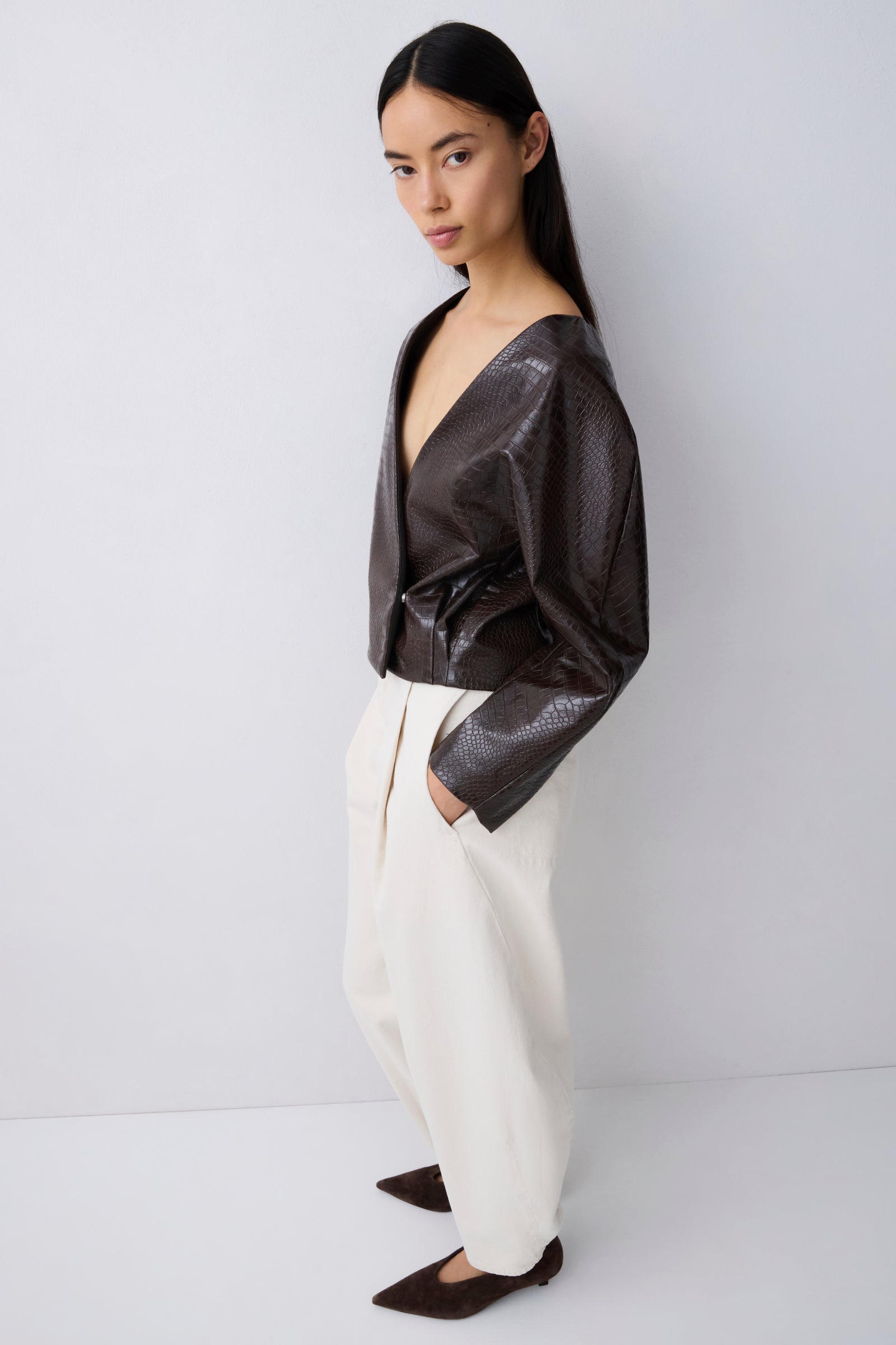 A model wearing a dark brown textured faux-leather wrap jacket and cream wide-leg trousers, standing against a light gray studio background.