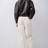 A model shown from the back wearing a dark brown textured faux-leather jacket with puffed sleeves, cream-colored wide-leg trousers, and dark brown pointed-toe heels, set against a plain light gray studio background.
