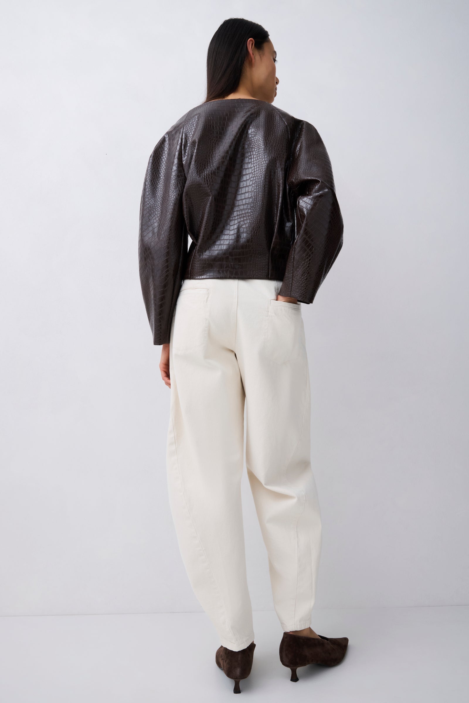 A model shown from the back wearing a dark brown textured faux-leather jacket with puffed sleeves, cream-colored wide-leg trousers, and dark brown pointed-toe heels, set against a plain light gray studio background.