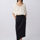 A model stands against a light gray studio background wearing a cream-colored crinkled blouse with ruffled sleeves and a black midi skirt.