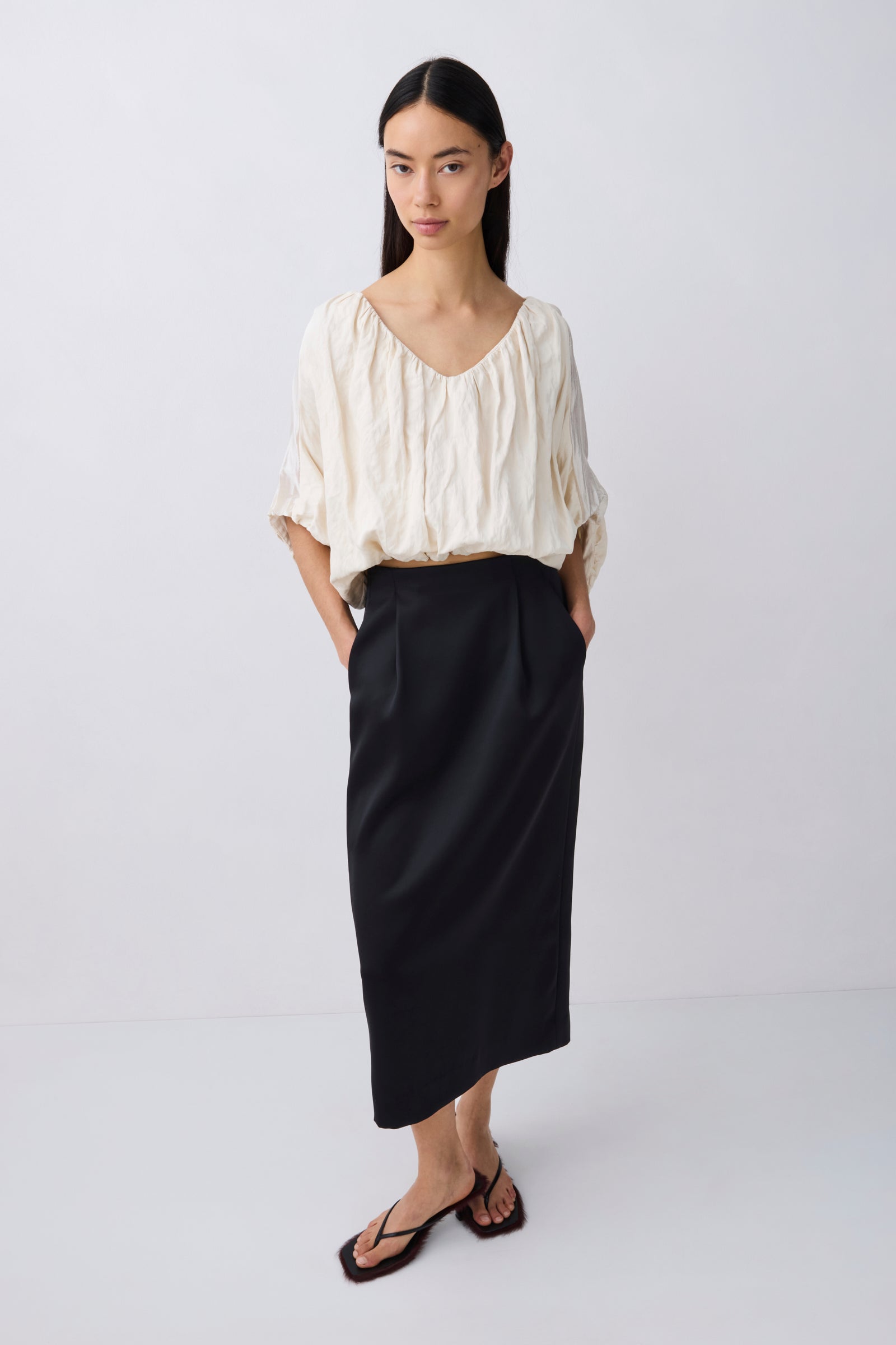 A model stands against a light gray studio background wearing a cream-colored crinkled blouse with ruffled sleeves and a black midi skirt.