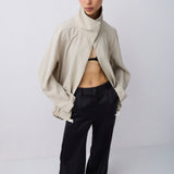 A model stands against a light gray studio background wearing a beige cropped jacket with a high folded collar, black wide-leg trousers, and a black bralette.
