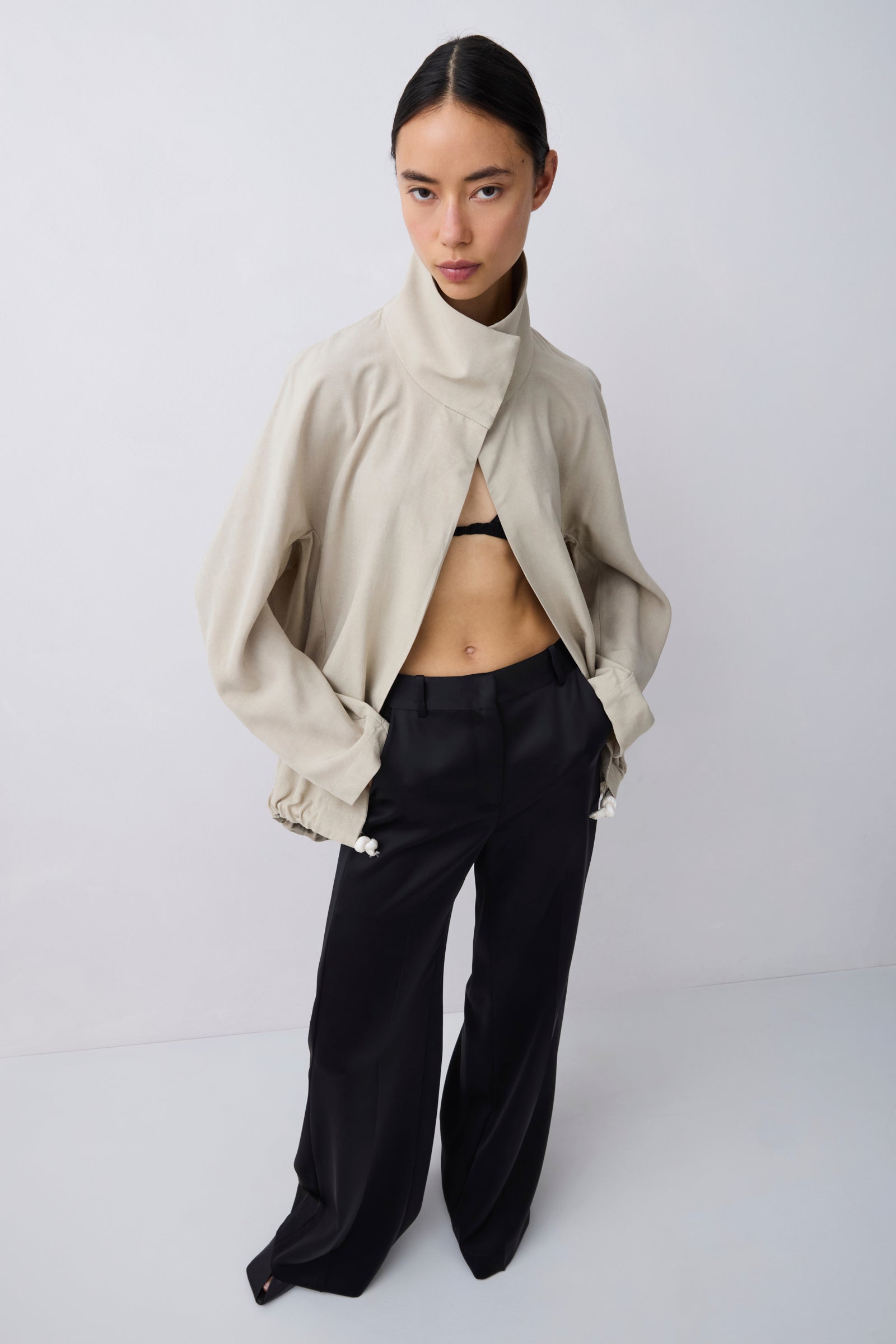 A model stands against a light gray studio background wearing a beige cropped jacket with a high folded collar, black wide-leg trousers, and a black bralette.