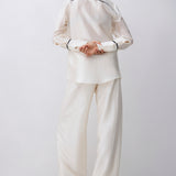 A model viewed from behind wearing a matching cream-colored two-piece pajama set with thin black piping details on the collar and cuffs, set against a plain light gray studio background.