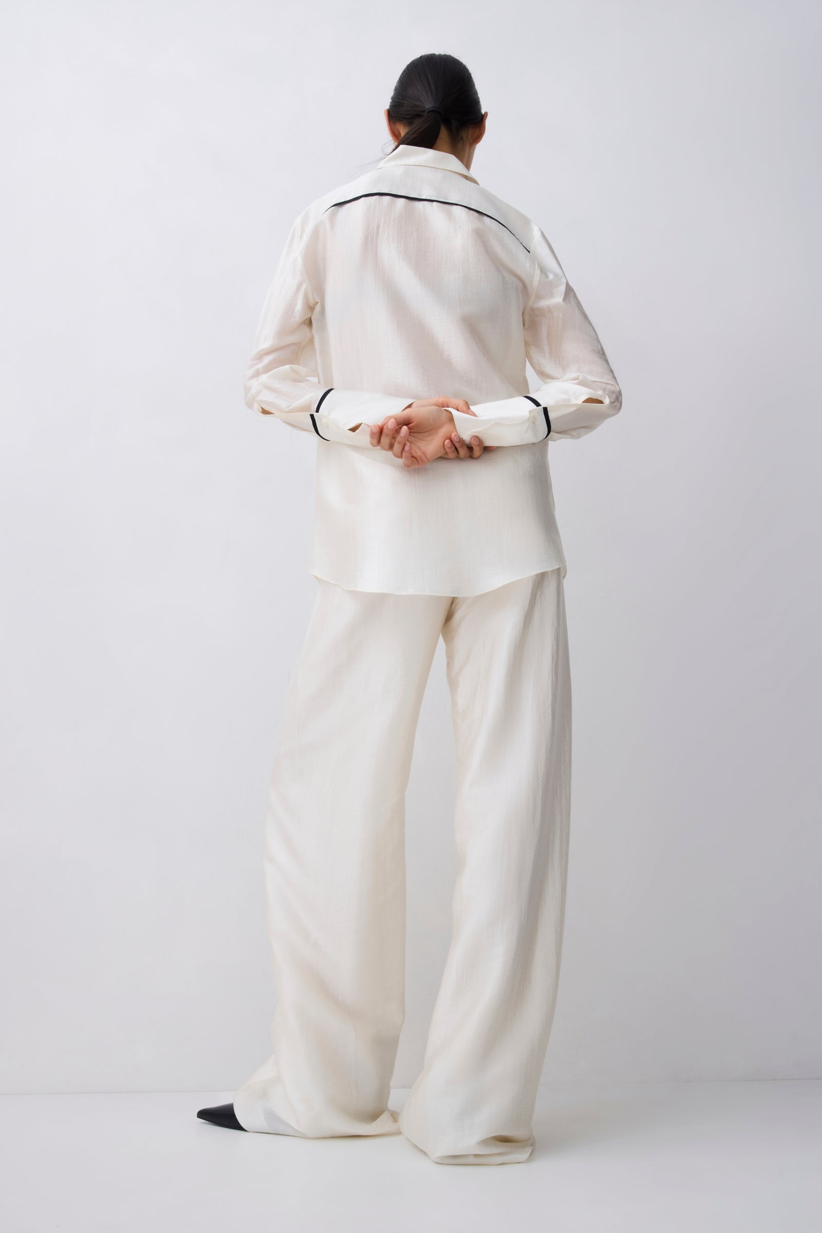 A model viewed from behind wearing a matching cream-colored two-piece pajama set with thin black piping details on the collar and cuffs, set against a plain light gray studio background.