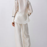 A model viewed from the back wearing a matching cream satin pajama set with black piping details on a light gray studio background.