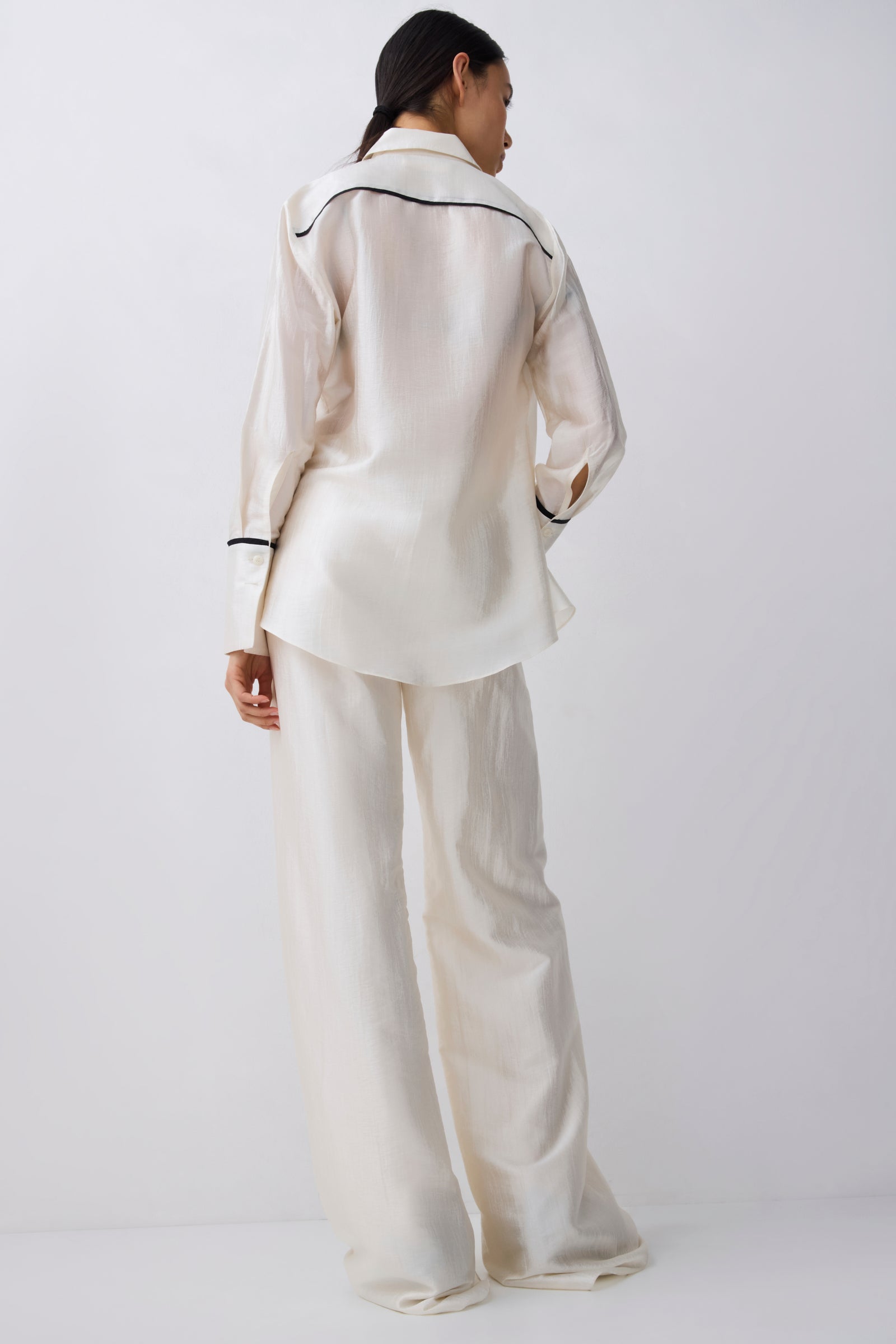 A model viewed from the back wearing a matching cream satin pajama set with black piping details on a light gray studio background.