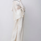 A model wearing a matching off-white satin pajama set with a long-sleeve button-up shirt and wide-leg pants, featuring thin black piping details, photographed against a light gray studio background.
