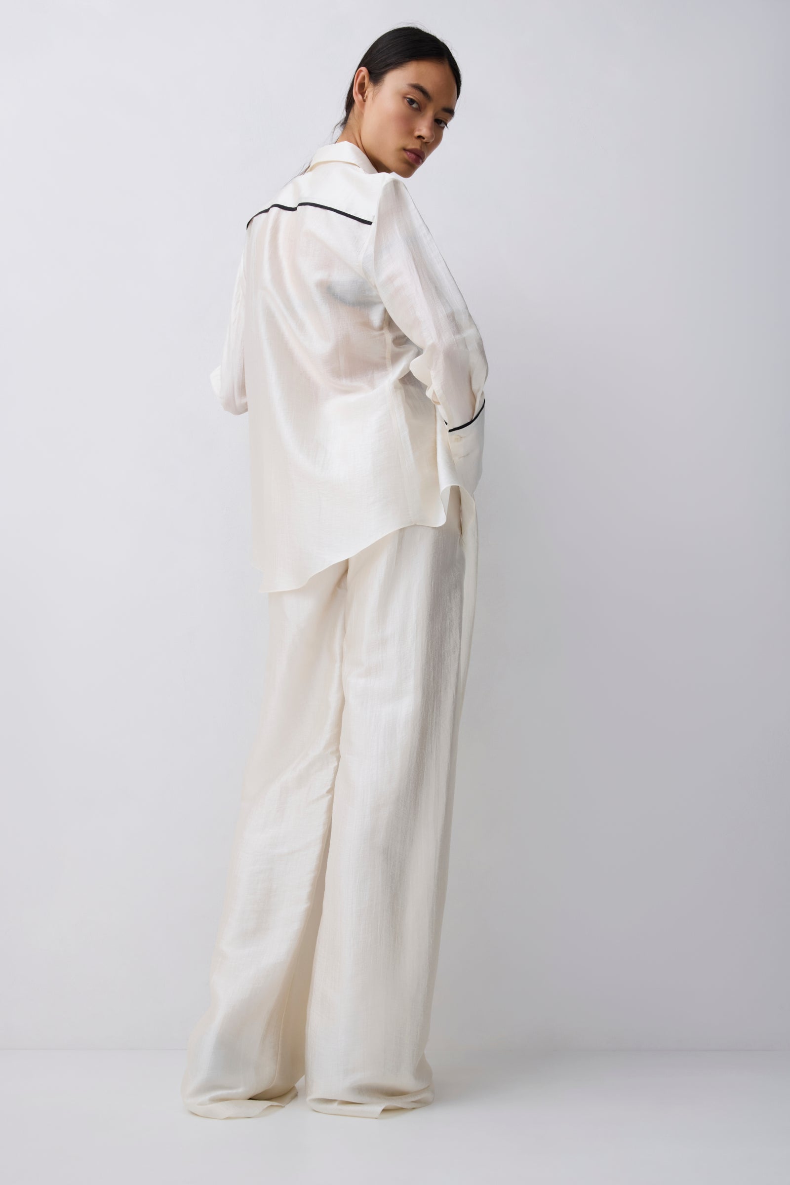 A model wearing a matching off-white satin pajama set with a long-sleeve button-up shirt and wide-leg pants, featuring thin black piping details, photographed against a light gray studio background.