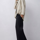 A model stands in a studio wearing a light beige oversized jacket with a high mock neck and voluminous sleeves, paired with wide-leg black trousers, against a light gray background.