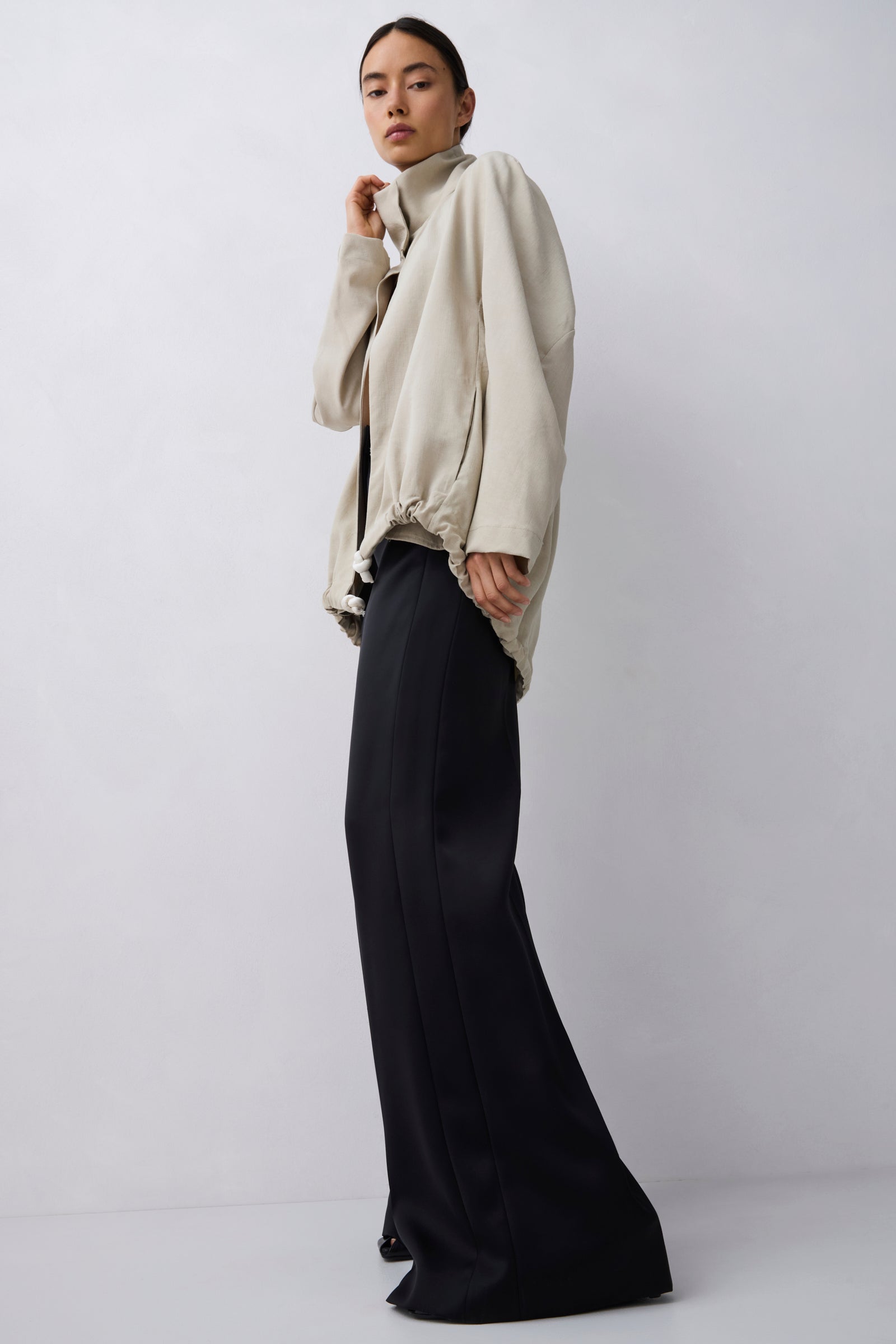 A model stands in a studio wearing a light beige oversized jacket with a high mock neck and voluminous sleeves, paired with wide-leg black trousers, against a light gray background.