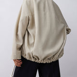 A model shown from the back wearing an oversized beige jacket with a gathered elastic hem and black wide-leg pants, set against a plain light gray studio background.