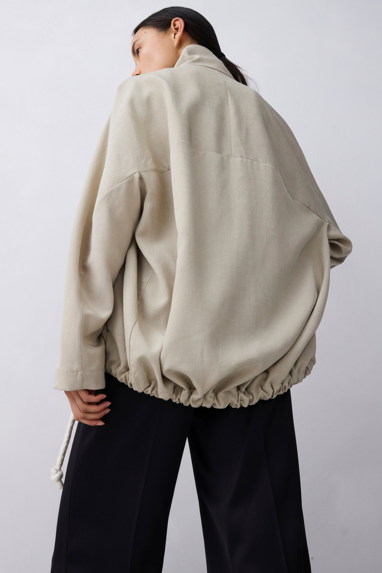 A model shown from the back wearing an oversized beige jacket with a gathered elastic hem and black wide-leg pants, set against a plain light gray studio background.