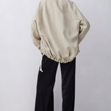 A model shown from the back wearing an oversized beige jacket with a drawstring hem and black wide-leg pants, set against a plain light gray studio background.