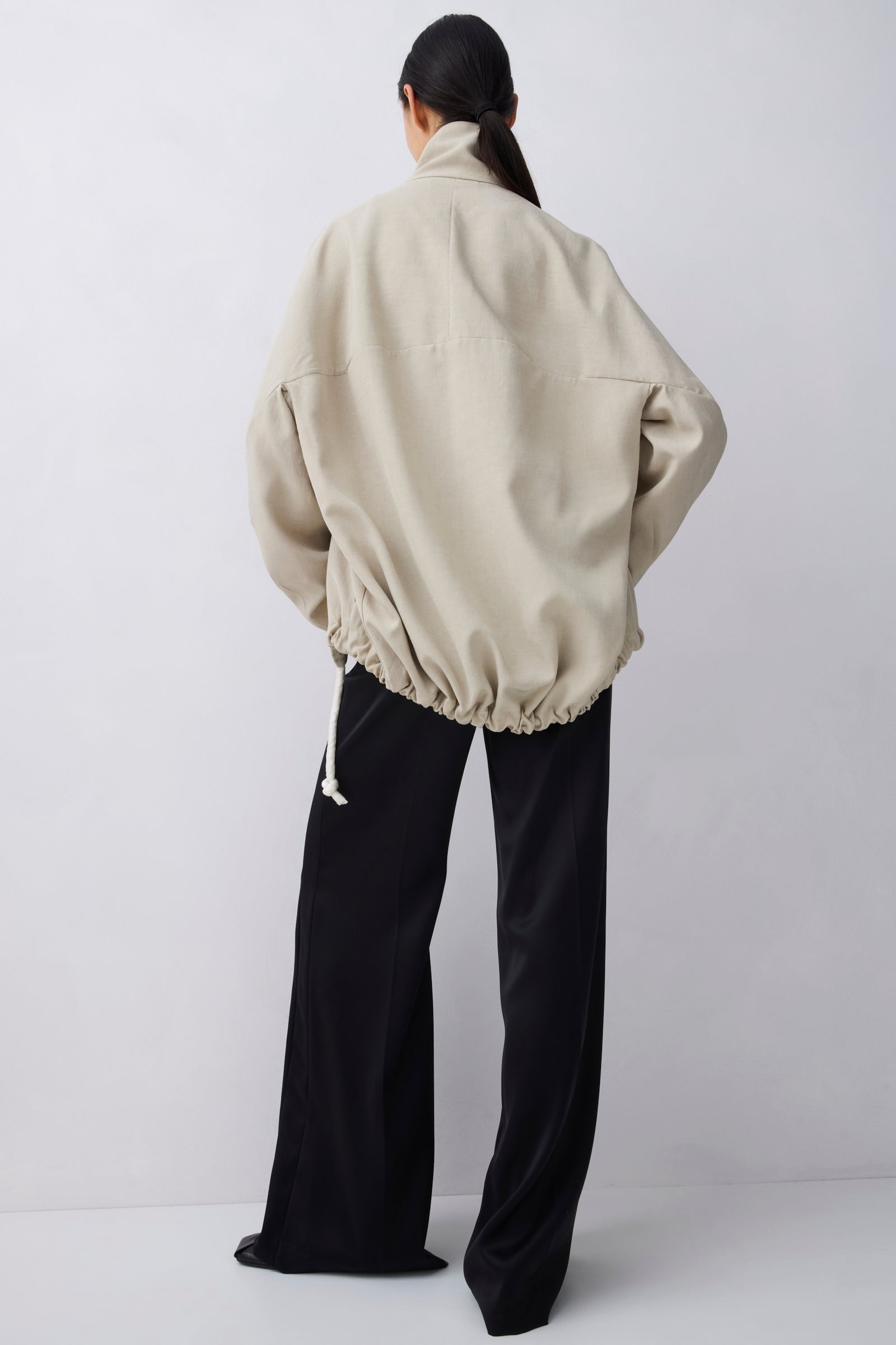A model shown from the back wearing an oversized beige jacket with a drawstring hem and black wide-leg pants, set against a plain light gray studio background.