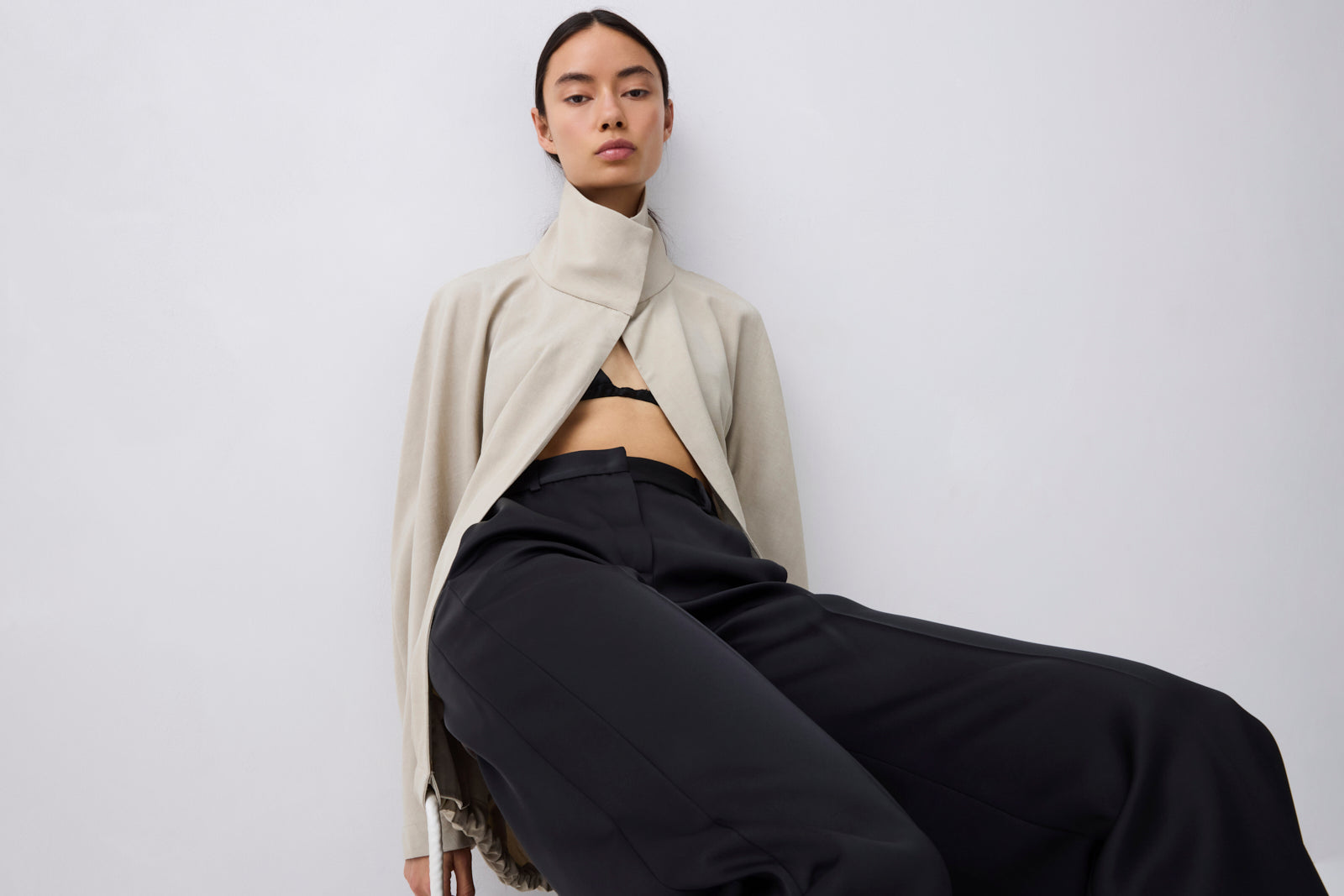 A model wearing a beige high-neck wrap jacket and black wide-leg trousers, seated against a plain light gray studio background.