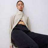 A model wearing a beige high-neck wrap jacket and black wide-leg trousers, seated against a plain light gray studio background.