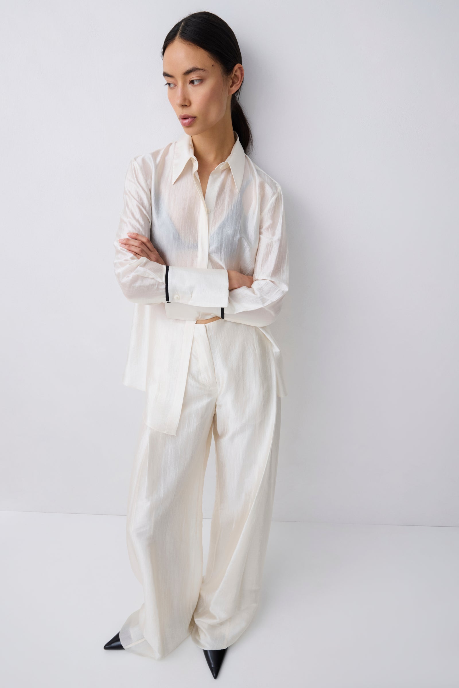 A model stands against a plain light gray studio wall wearing an oversized white long-sleeve button-up shirt and matching wide-leg trousers, with black pointed shoes.