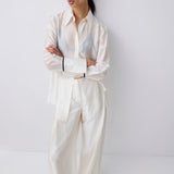 A model stands against a plain light gray studio wall wearing an oversized white long-sleeve button-up shirt and matching wide-leg trousers, with black pointed shoes.