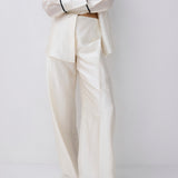 A model wearing a matching cream-colored two-piece suit consisting of a long-sleeve button-down shirt and wide-leg trousers, styled with black pointed shoes against a plain light gray background.
