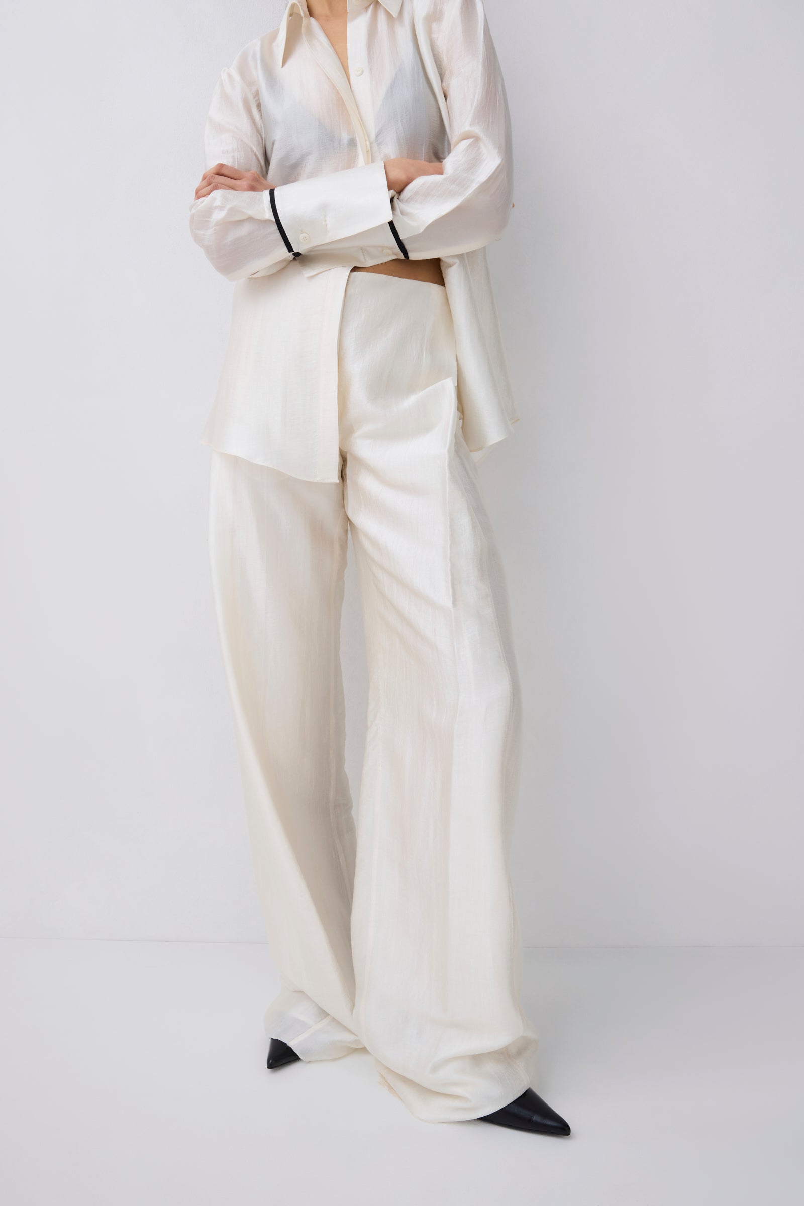 A model wearing a matching cream-colored two-piece suit consisting of a long-sleeve button-down shirt and wide-leg trousers, styled with black pointed shoes against a plain light gray background.
