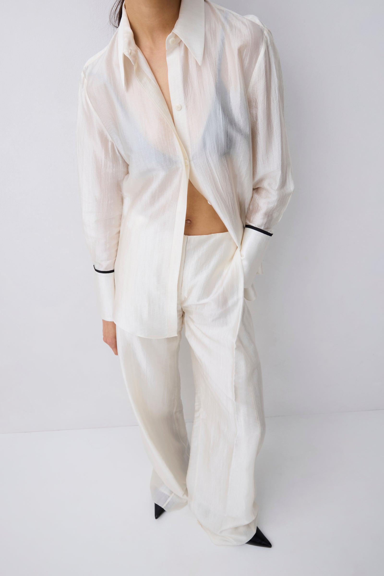 A model wearing a matching off-white two-piece lounge set consisting of a long-sleeve button-up shirt and wide-leg trousers, styled with black pointed shoes against a light gray studio background.