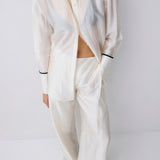 A model wearing a matching off-white two-piece lounge set consisting of a long-sleeve button-up shirt and wide-leg trousers, styled with black pointed shoes against a light gray studio background.