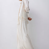 A model wearing a flowing, oversized white or cream satin-like two-piece outfit with draped layers and wide-leg pants, posed against a plain light gray studio background.