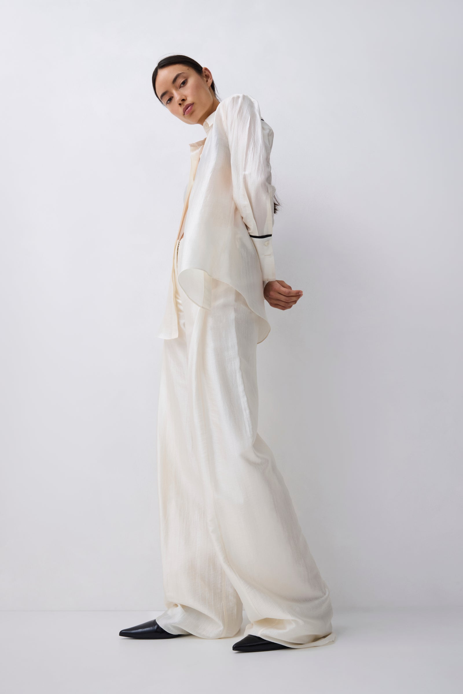 A model wearing a flowing, oversized white or cream satin-like two-piece outfit with draped layers and wide-leg pants, posed against a plain light gray studio background.