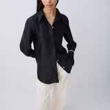 A woman modeling a black long-sleeve button-up shirt with white cuff stripes, paired with cream-colored pants and black sandals, against a light gray studio background.