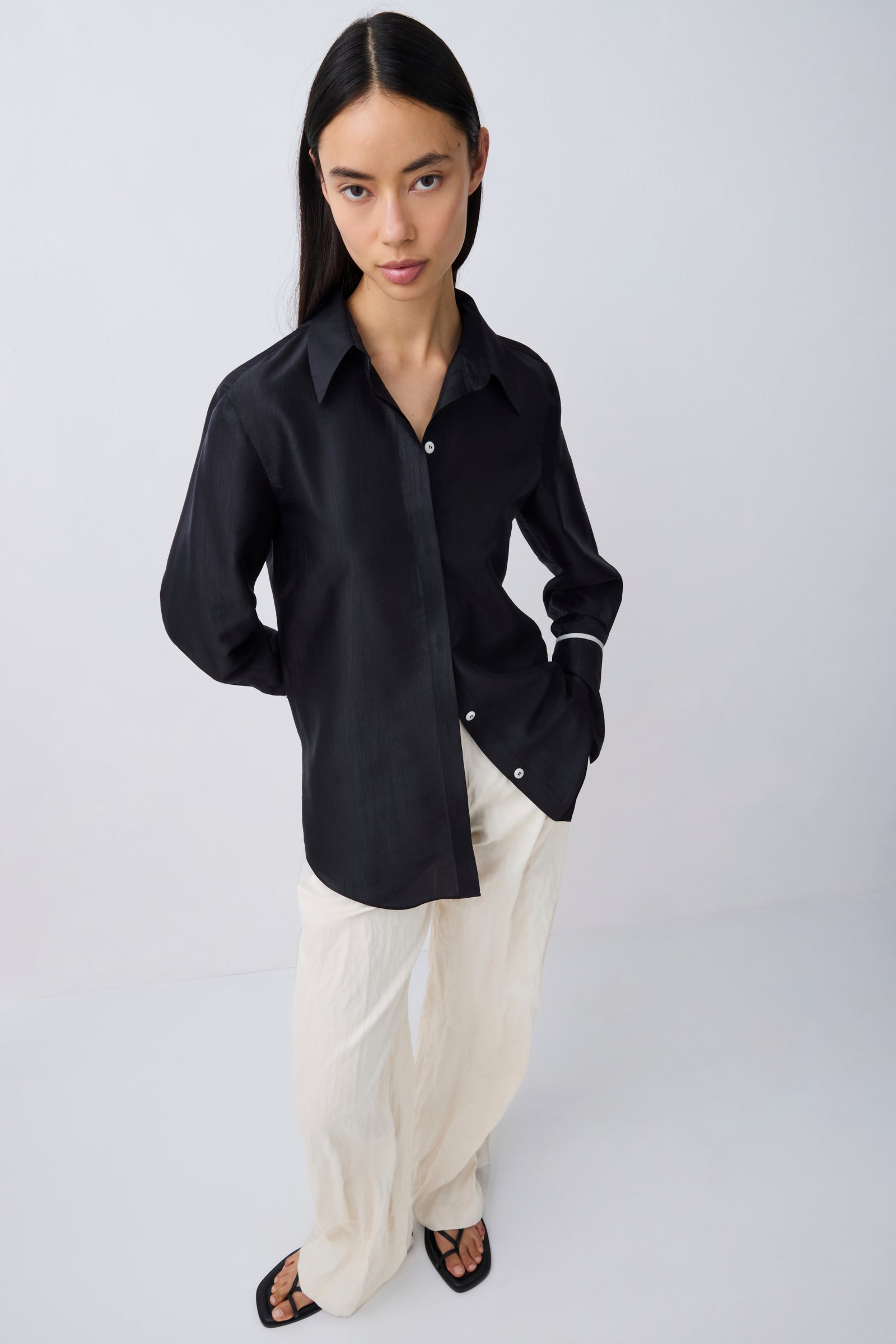A woman modeling a black long-sleeve button-up shirt with white cuff stripes, paired with cream-colored pants and black sandals, against a light gray studio background.