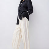 A model wearing a black satin long-sleeve button-up shirt and cream wide-leg trousers, paired with black sandals, against a plain light gray studio background.