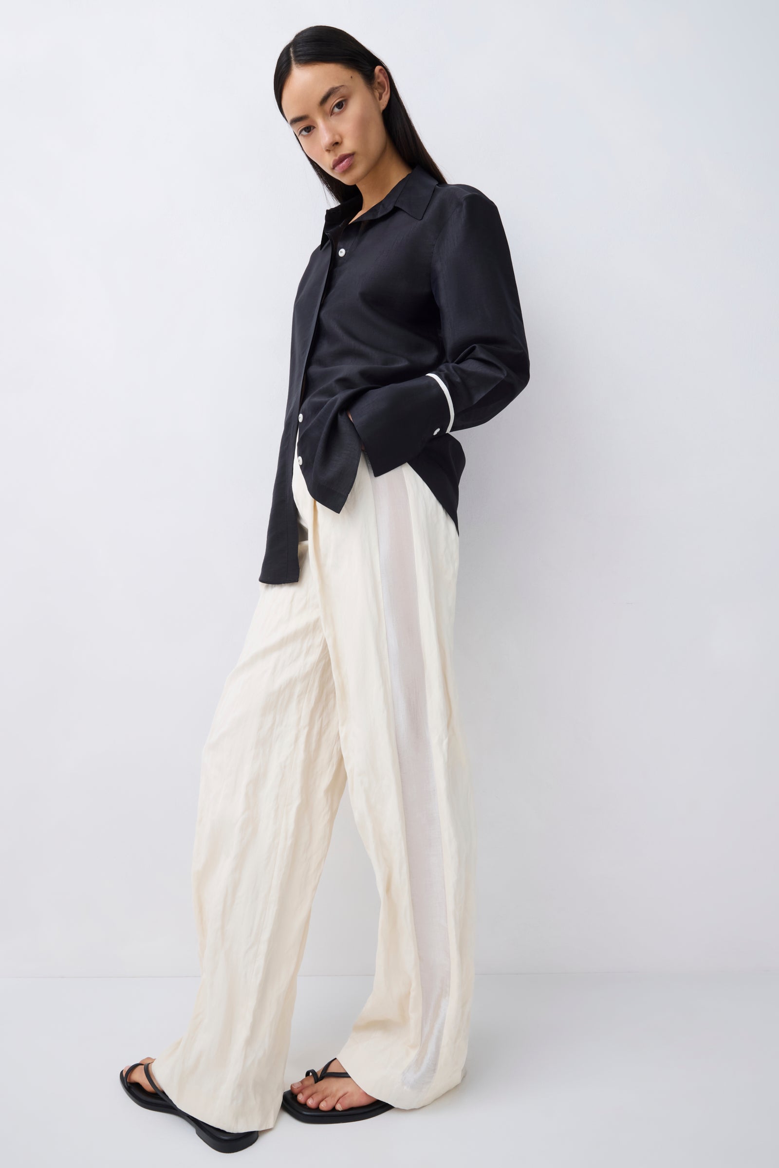 A model wearing a black satin long-sleeve button-up shirt and cream wide-leg trousers, paired with black sandals, against a plain light gray studio background.