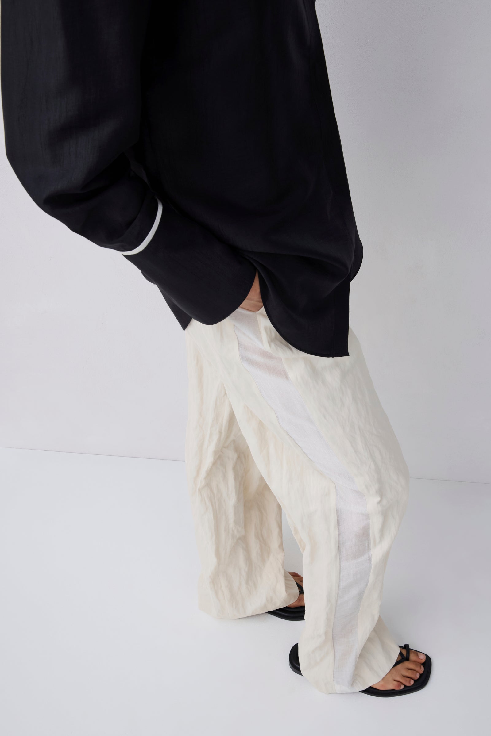 A person wearing loose, oversized off-white textured pants with a black top and black sandals, shown from the chest down against a plain light gray studio background.