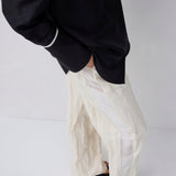 A person wearing loose, oversized off-white textured pants with a black top and black sandals, shown from the chest down against a plain light gray studio background.