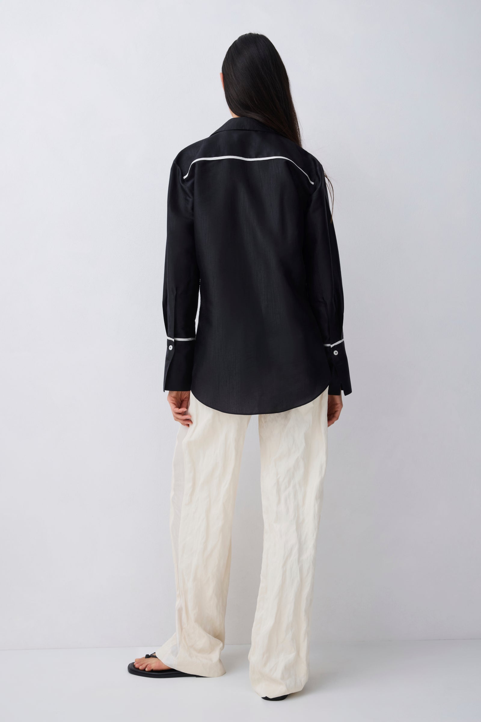A model shown from the back wearing a black long-sleeve western-style shirt with white contrast stitching and cream wide-leg trousers, standing against a light gray studio background.