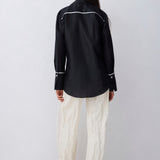 A model shown from the back wearing a black long-sleeve western-style shirt with white contrast stitching and cream wide-leg trousers, standing against a light gray studio background.