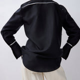 A model shown from the back wearing a black long-sleeve shirt with thin white piping details along the collar and cuffs, paired with cream-colored pants, against a light gray background.