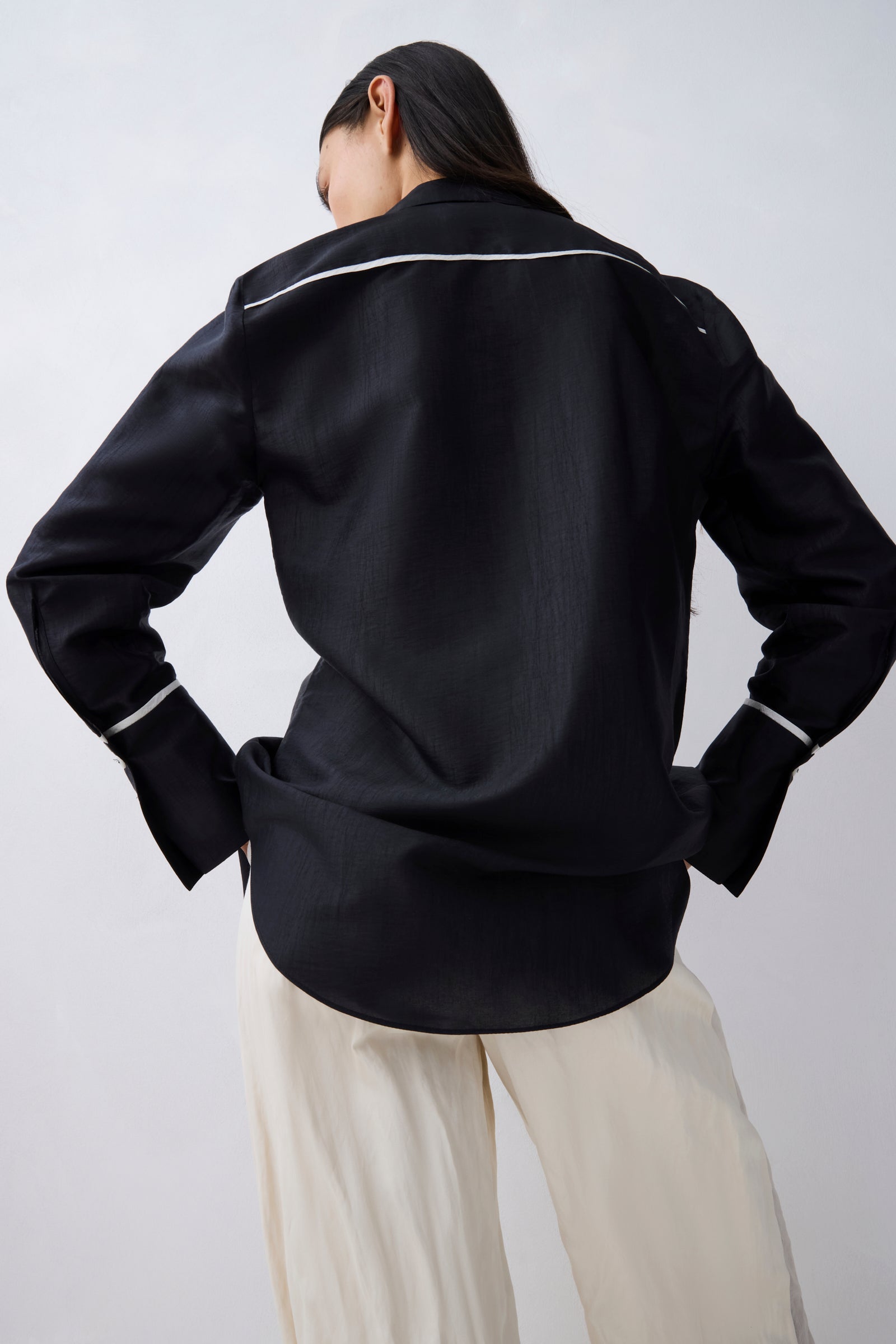 A model shown from the back wearing a black long-sleeve shirt with thin white piping details along the collar and cuffs, paired with cream-colored pants, against a light gray background.