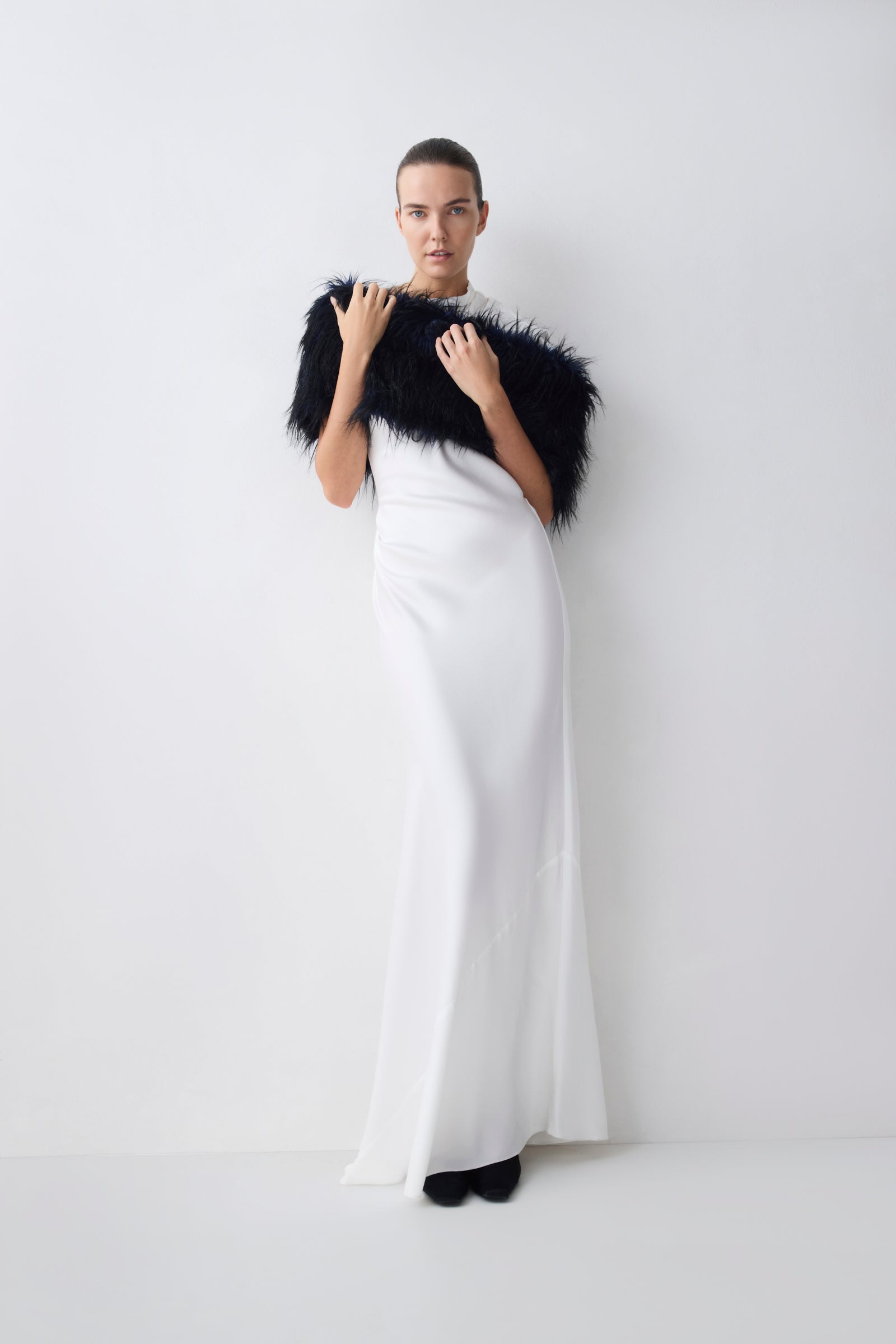 Woman wearing a navy fur shrug over a white dress on a plain background